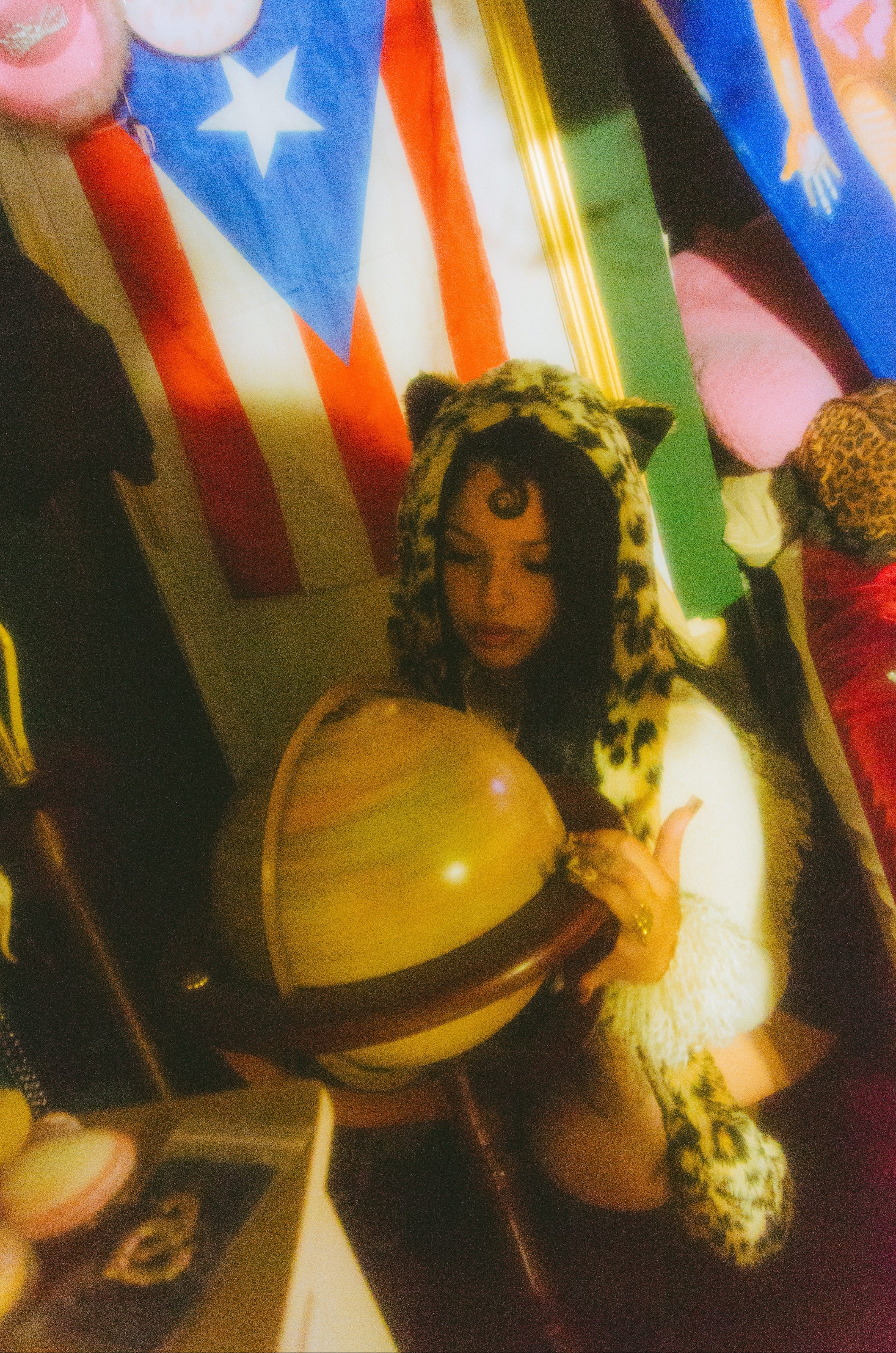 A person wearing a leopard-print cat-ear hooded outfit kneels behind a large yellow globe, holding it in a dimly lit room with colorful flags and plush items in the background.