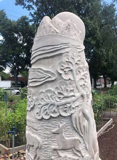 A large carved stone garden sculpture featuring a mountain, waves, leaves, deer, and a deer head, photographed outdoors in a garden setting with trees and plants in the background.